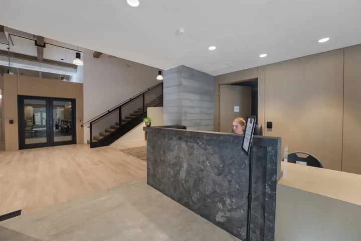 Modern reception area featuring light wood-finish flooring, a contemporary staircase with black railing and glass panels, and a stone-clad reception desk