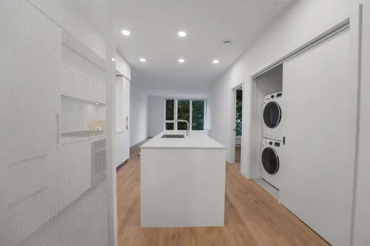 Modern kitchen featuring a central island with an integrated sink, light wood-finish flooring, recessed lighting, floor-to-ceiling windows, and a built-in stackable washer and dryer unit behind a sliding door