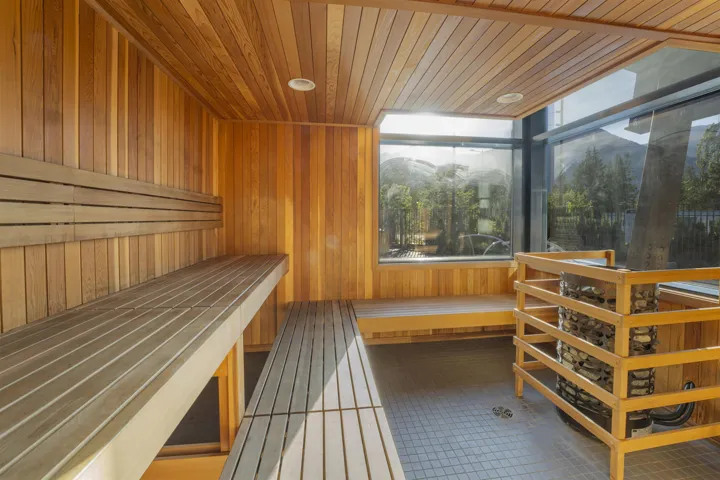 Wood-paneled sauna featuring a built-in multi-level bench, natural wood wall and ceiling finishes, a heated rock element with a wood enclosure, a large picture window, and square-tiled flooring