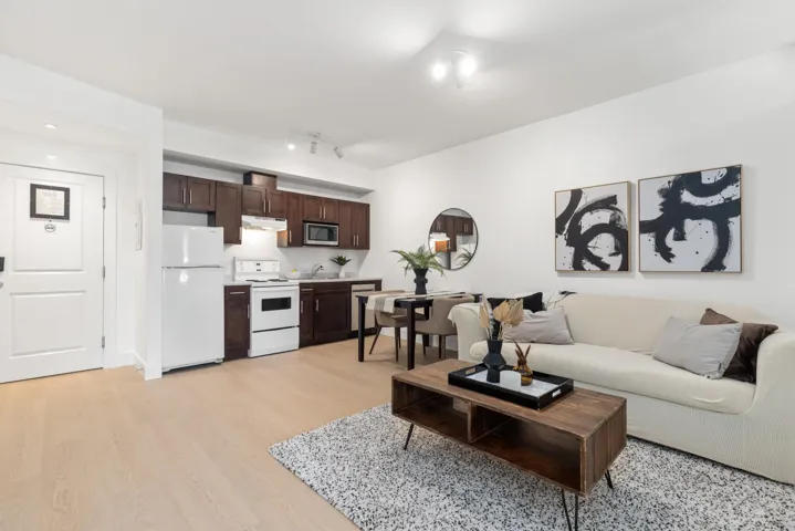 Open-concept living area featuring light wood-finish flooring, white walls, and a modern kitchen with dark wood cabinetry and white appliances