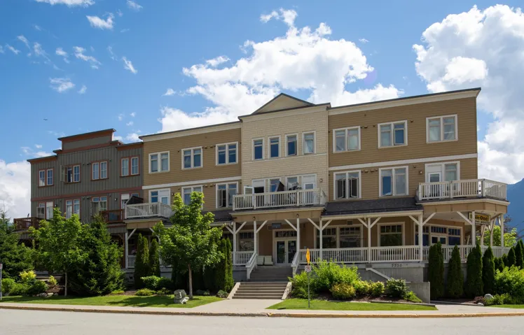 Multi-story building featuring balconies with white railings, a prominent main entrance with steps, and a landscaped frontage with mature trees and shrubs