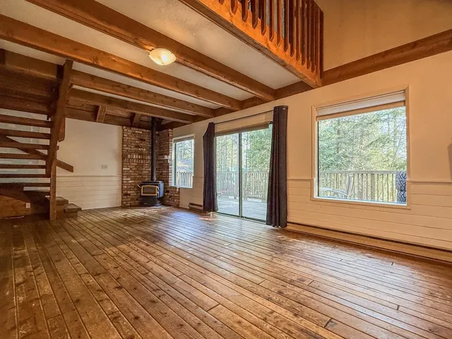 Unfurnished living room with a wood stove, a wainscoted wall, beam ceiling, wood-type flooring, and wood walls