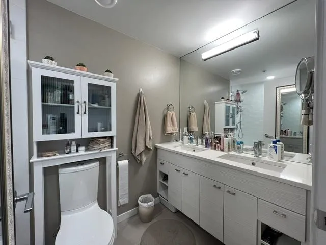 Bathroom featuring a dual vanity with integrated sinks, a full-wall mirror, and an over-toilet storage cabinet with fluted glass doors