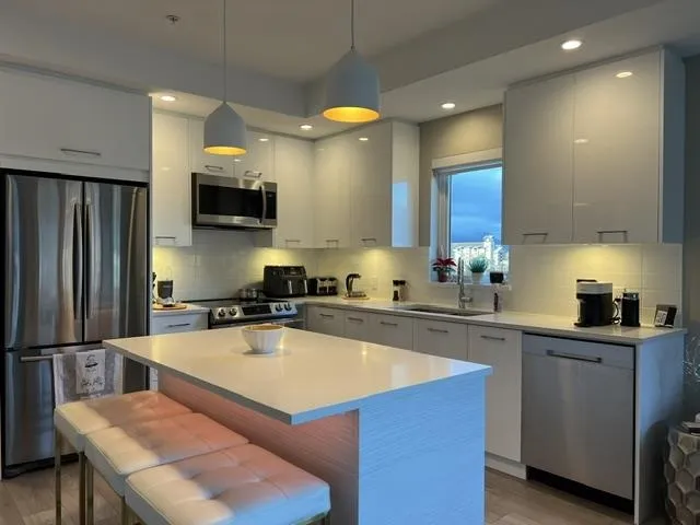 Modern kitchen featuring a central island with countertop seating, glossy white cabinetry, stainless steel appliances including a refrigerator, microwave, and dishwasher, and a white subway tile backsplash