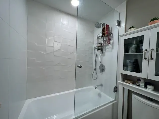 White bathtub and shower enclosure featuring a clear glass panel, textured white shower wall tile, and built-in white cabinetry with fluted glass doors