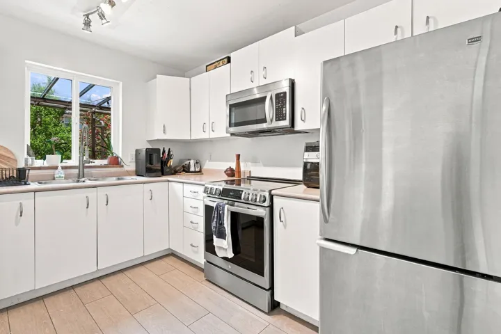 Modern kitchen featuring white cabinetry, stainless steel appliances, light-toned countertops, wood-finish flooring, and a large window above the sink