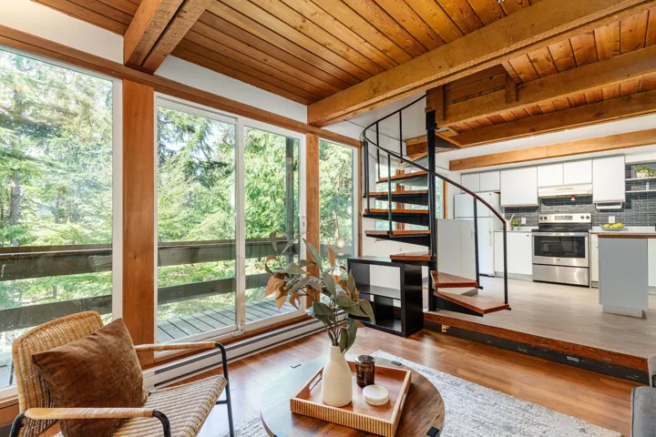 Wood-beam ceiling with exposed joists, full-height windows, spiral staircase, wood-finish flooring, and a kitchen featuring white cabinetry, subway tile backsplash, and stainless steel appliances