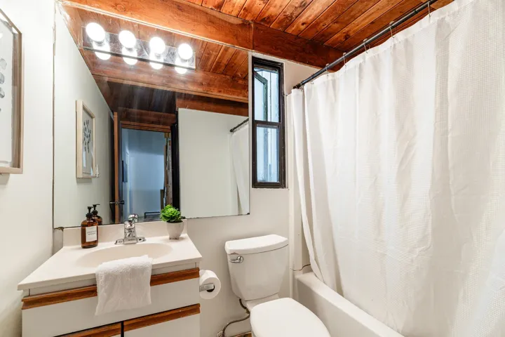 Wood-paneled ceiling, integrated vanity with single basin sink, wall-mounted mirror with overhead lighting, and a full bathtub with shower