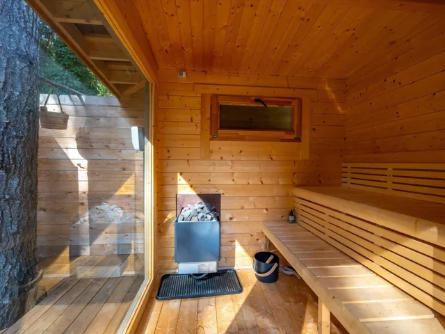 Wood-paneled sauna featuring a built-in multi-level bench, a rock-filled heater, and a large transparent panel providing an exterior view