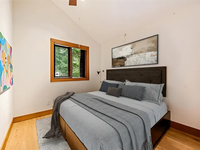 Bedroom with vaulted ceilings, natural wood-finish flooring, and a large window with wood blinds