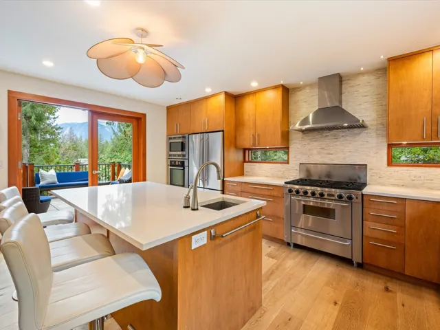 Modern kitchen featuring wood-finish flooring, light wood cabinetry, stainless steel appliances, a center island with integrated sink, and a tiled backsplash