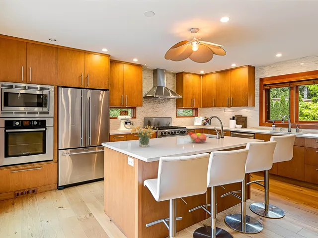Kitchen featuring wood cabinetry, stainless steel appliances including a built-in oven and microwave, a center island with a white countertop and integrated sink, and light wood-finish flooring