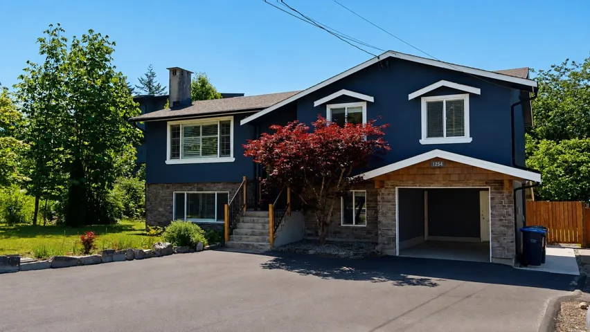 Exterior featuring a dark blue facade, white trim windows, a stone-fronted garage, a shingle-accented entryway, and mature landscaping