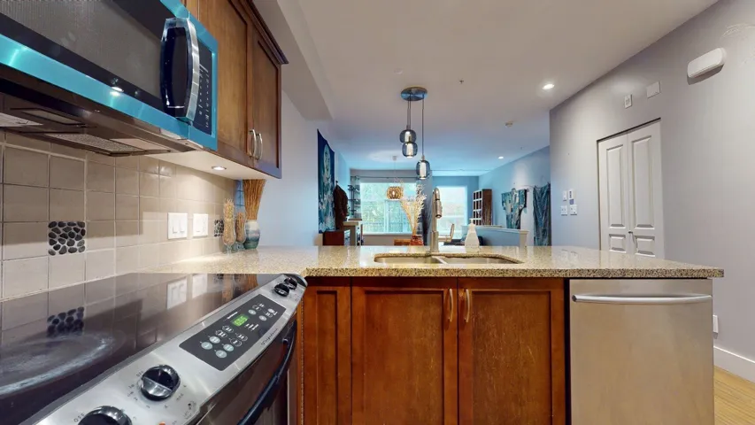 Kitchen with brown cabinets, stainless steel appliances, hanging light fixtures, a peninsula, and light stone counters