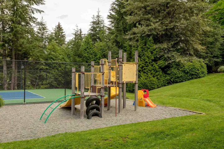 Outdoor recreation area featuring a playground with slides and climbing elements, a fenced tennis court with a blue and green surface, and surrounding evergreen trees