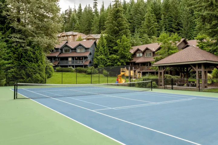 Full-size tennis court featuring a blue playing surface with white line markings, surrounded by a green perimeter