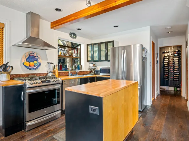 Kitchen featuring wood counters, appliances with stainless steel finishes, wall chimney range hood, beam ceiling, and dark wood-style flooring