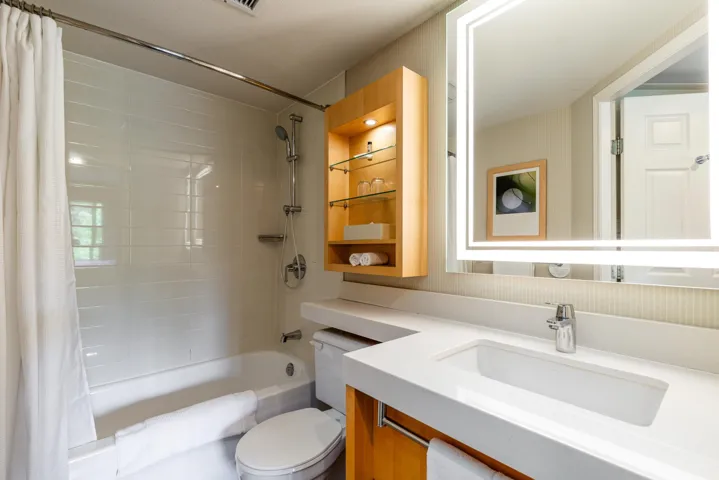 Bathroom featuring an illuminated mirror, a white rectangular sink, a single-handle chrome faucet, a built-in wooden cabinet with glass shelving, and a combination tub and shower with white subway tile surround