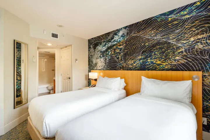 Bedroom featuring two beds with light wood headboards, a distinctive accent wall with abstract patterning, and a wall-mounted full-length mirror
