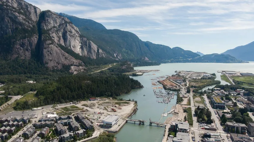 Coastal cityscape with a marina, surrounded by towering mountains and extensive forested areas