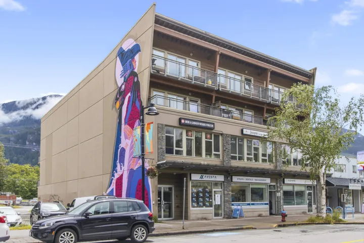 Modern mixed-use building featuring a distinct angled facade with a large-scale mural, ground-level retail spaces, upper-level residential balconies with glass railings, natural wood accents, and stone-clad lower levels