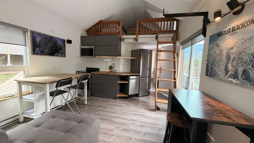 Open-concept living area featuring wood-finish flooring, a contemporary kitchen with dark cabinetry and stainless steel appliances, and a built-in loft with a wooden ladder