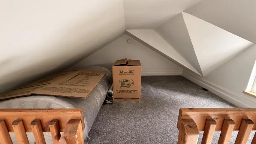 Attic space featuring sloped ceilings, light gray carpet, white baseboards, and a window providing natural illumination