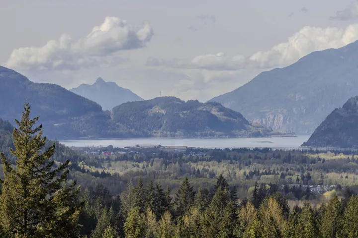 Expansive mountain and water scenery featuring a body of water framed by multiple mountain ranges, a verdant forest, and distant structures