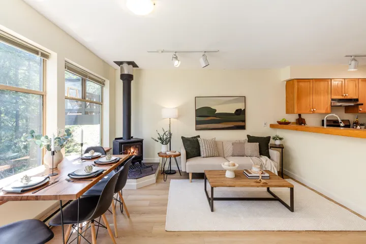 Open-concept living area with wood-finish flooring, featuring a dining space with large windows, a wood-burning stove on a tiled hearth, and track lighting