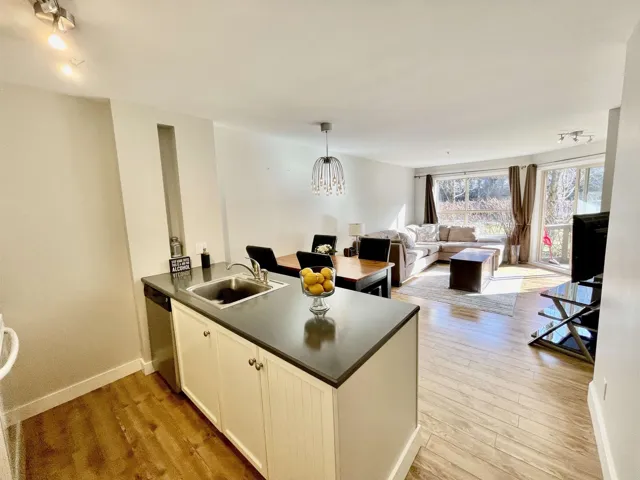 Open-concept living area with wood-finish flooring, a kitchen island featuring a stainless steel sink, and a dining area with a contemporary chandelier