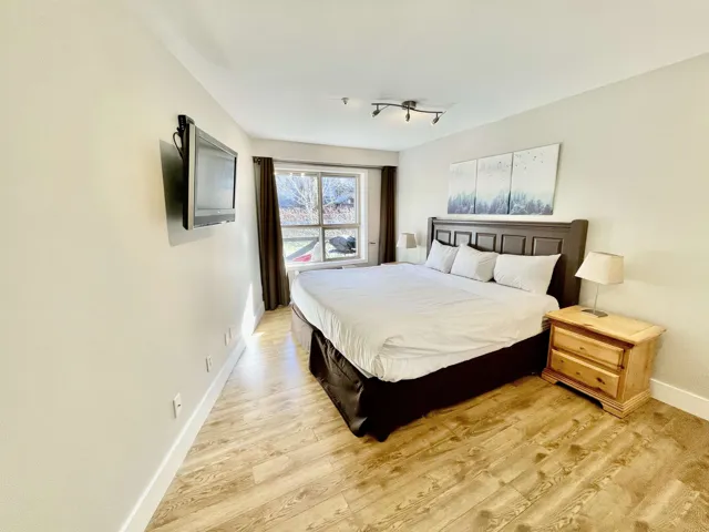 Spacious bedroom featuring light-toned walls, wood-finish flooring, a contemporary track lighting fixture, and a large window with privacy drapes