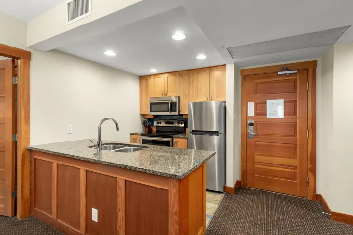 Kitchen area featuring a granite-topped island with an undermount double sink, stainless steel appliances, wood cabinetry, and recessed lighting