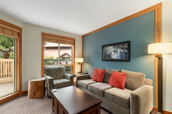 Living area featuring a teal accent wall with wood-finish trim, large window with natural wood-finish blinds, light-toned carpeting, and a glass sliding door providing outdoor access