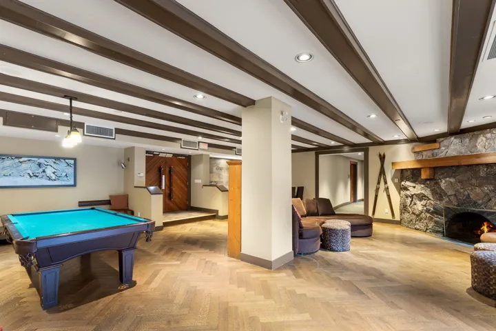 Spacious recreational room featuring a herringbone wood-finish floor, exposed beam ceiling, stone fireplace with a wood mantle, and double wood entry doors
