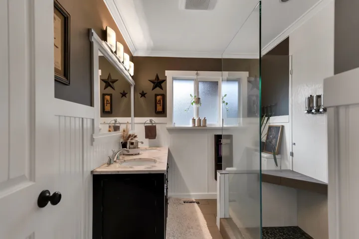 Bathroom featuring a dark wood vanity with a light-toned countertop, a glass-enclosed shower with a built-in bench, white wainscoting, and crown molding