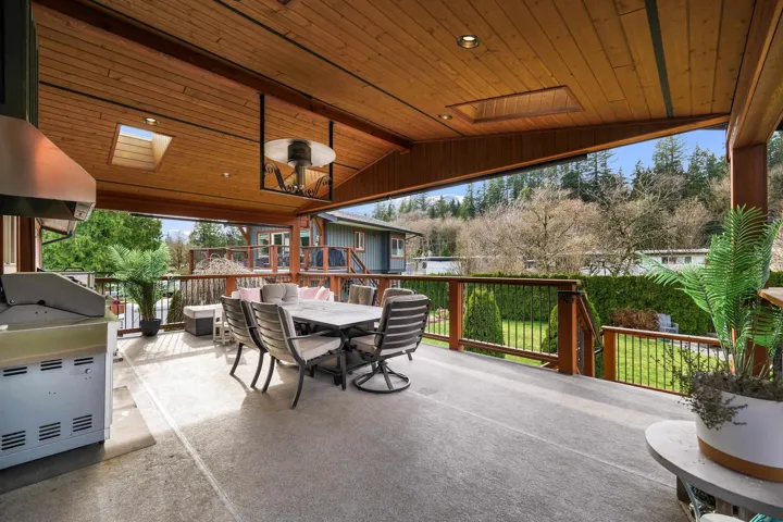 Expansive covered deck featuring a wood-panel ceiling with recessed lighting and skylights, outdoor kitchen with built-in grill, and wood railing with black iron spindles