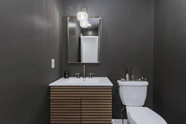 Textured wood-finish vanity with a rectangular sink, white countertop, gold-toned hardware, wall-mounted light fixture, and a framed mirror