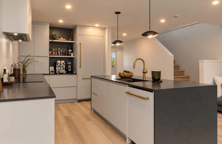 Kitchen featuring a center island with a dark countertop and integrated sink, flat-panel cabinetry, wood-finish flooring, brass-tone hardware, and pendant lighting