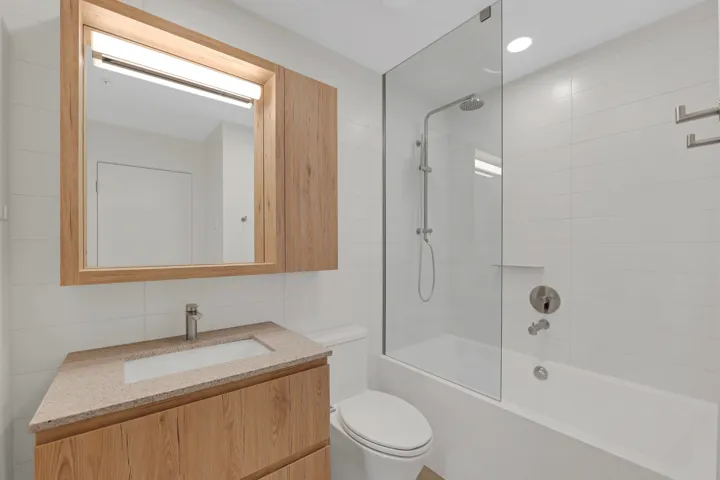 Bathroom featuring a wood-finish vanity with integrated sink, a speckled countertop, framed mirror with integrated lighting, a glass-enclosed bathtub with shower system, and white tiled walls