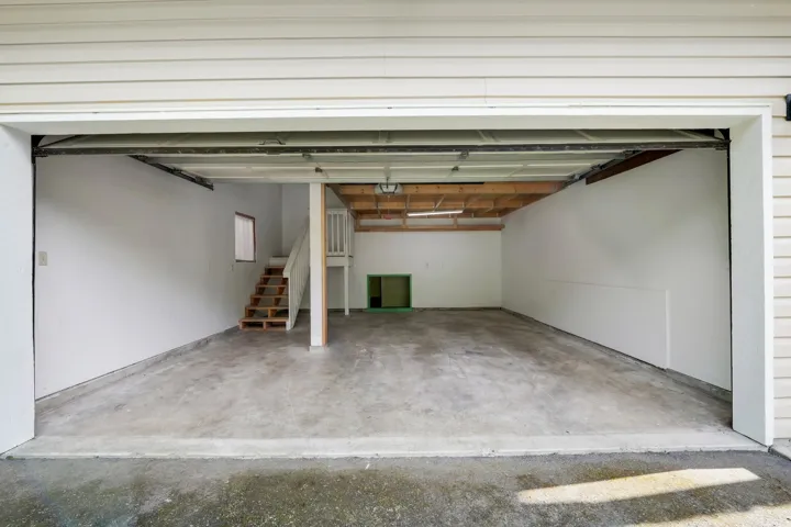Spacious garage featuring a concrete floor, white painted walls, exposed ceiling joists, and a wooden staircase providing access to an upper level