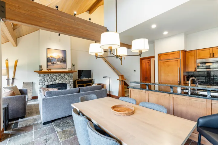 Vaulted ceilings with exposed wood beams over a great room featuring stone flooring, a river rock fireplace, and an open-concept kitchen with wood cabinetry and stainless steel appliances