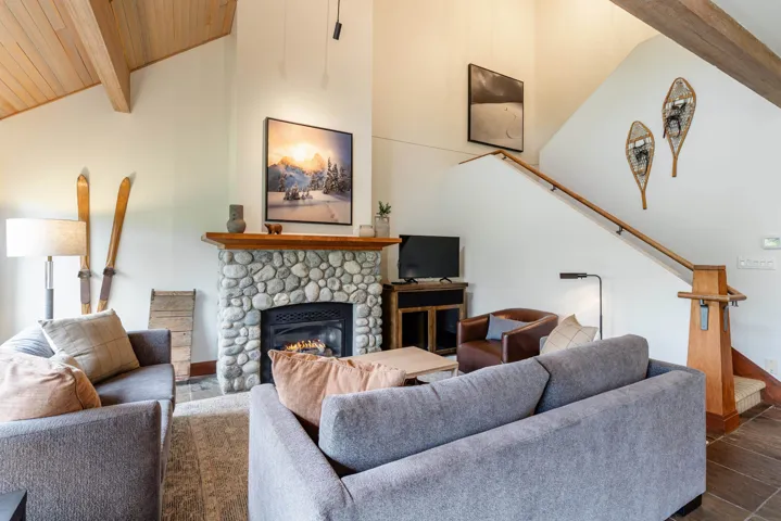 Living area featuring a river stone fireplace with a wood mantel, vaulted wood-paneled ceiling, exposed wood beams, and a natural wood staircase with a matching railing