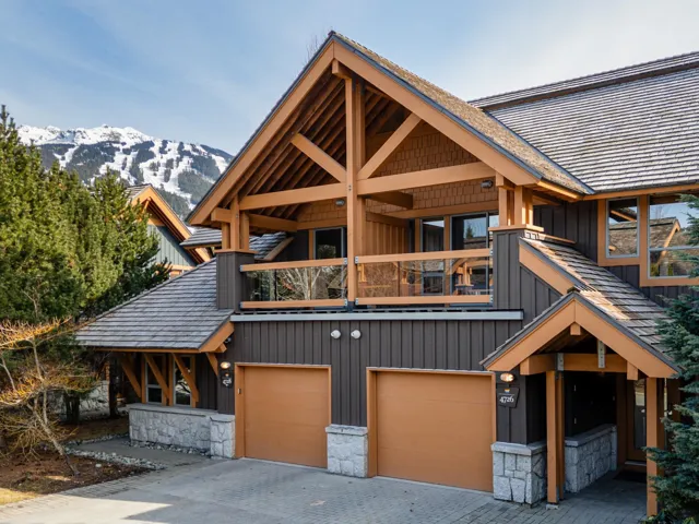 Timber frame exterior with cedar shake roofing, stone accents, two garage doors, and an upper-level balcony with glass railing