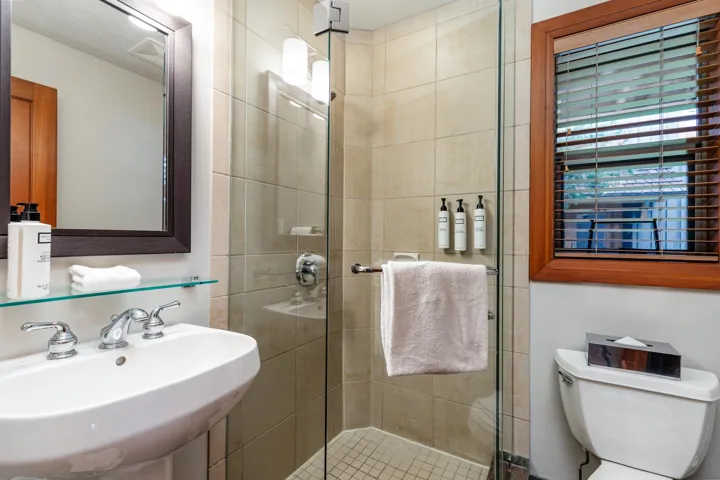 Pedestal sink with chrome fixtures and framed mirror, glass-enclosed shower with tiled surround, and toilet with wood-framed window
