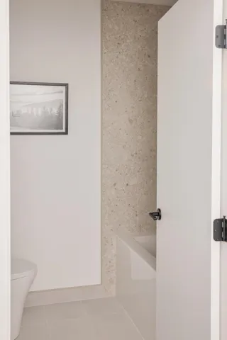 Bathroom featuring a built-in tub with a light-toned stone surround, a white toilet, and large format tile flooring