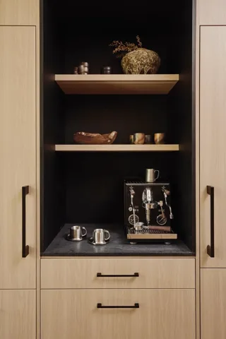 Built-in coffee bar featuring light wood-finish cabinetry, matte black hardware, floating wooden shelves, and a dark countertop
