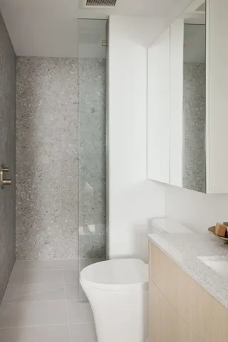 Bathroom featuring a glass-enclosed shower with stone-finish wall tile, a freestanding toilet, a light wood-finish vanity with a stone-finish countertop, and white overhead cabinetry with integrated mirrors