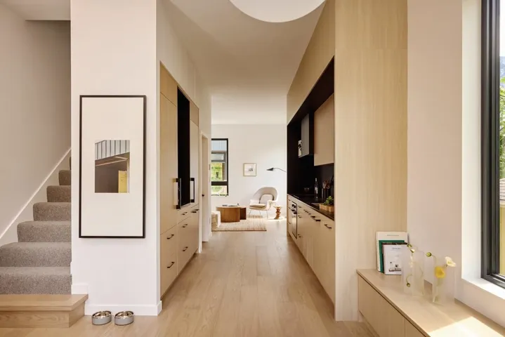 Open-concept interior featuring light wood-finish flooring, a contemporary staircase with carpeted treads, and an integrated kitchen with light wood cabinetry and dark countertops