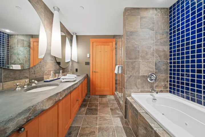 Full bathroom featuring a tub with jets, a stall shower, double vanity, tile walls, and stone tile flooring