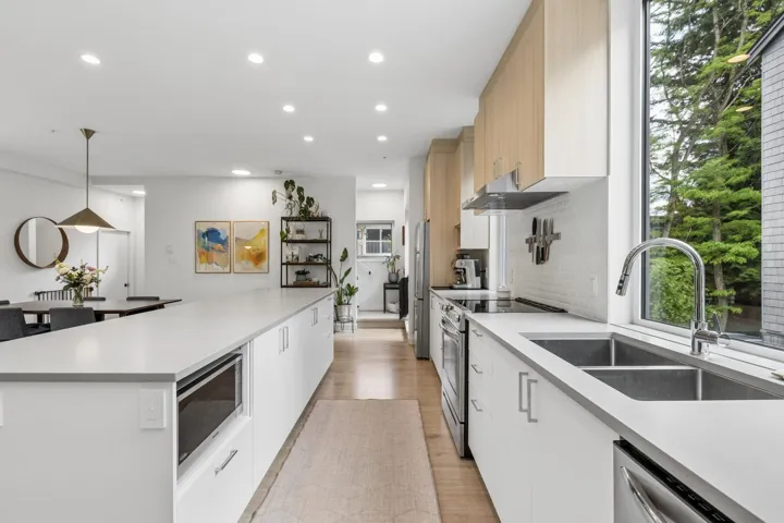 Contemporary kitchen featuring a long island with a built-in microwave, light wood-finish cabinetry, stainless steel appliances, a double basin sink, and a large window overlooking exterior trees